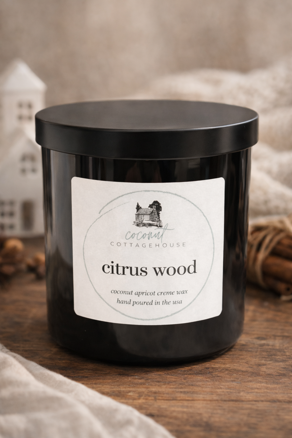 A black candle labeled "citrus wood" from the "Cocoon Cottage" brand, sitting on a wooden surface with a blurred background.
