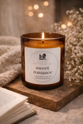 A brown glass candle labeled "Sweet Romance" sits on a wooden surface, with a blurred background featuring a book and a bouquet of white flowers.