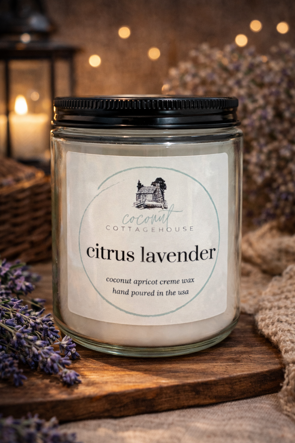 A glass jar candle labeled "Citrus Lavender" from "Cocot Cottage" sits on a wooden surface, surrounded by purple flowers and a lantern in the background.