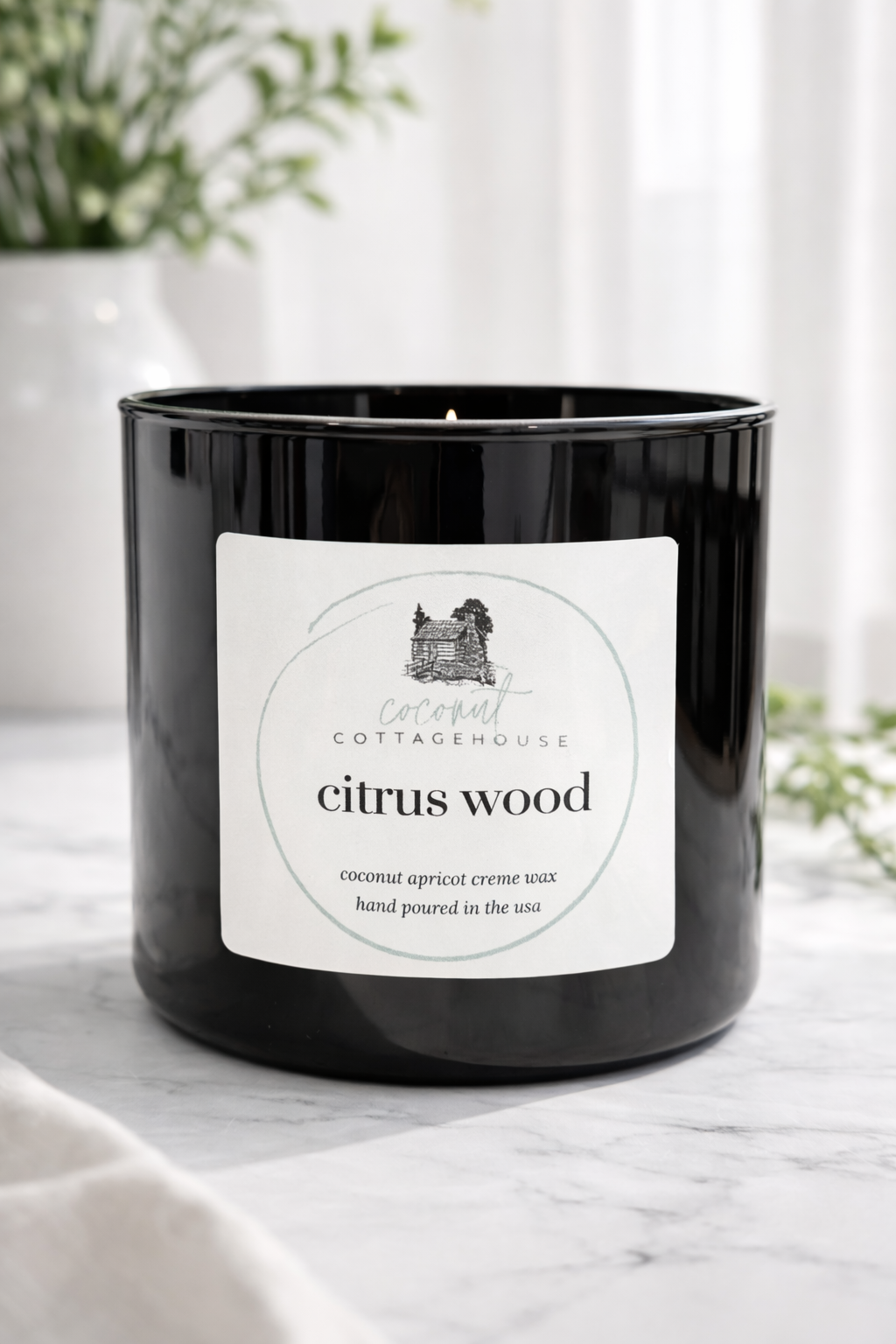 A black candle labeled "Citrus Wood" from the "Cottage Cottage" brand, sitting on a marble surface with a white vase of greenery in the background.