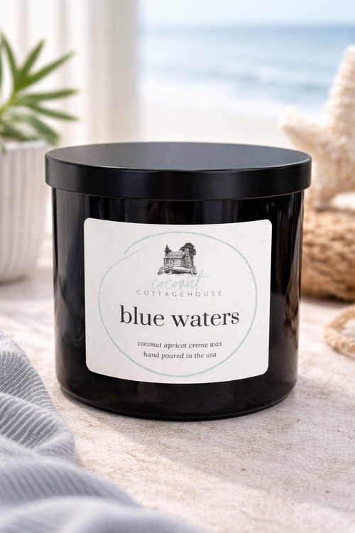 A black candle labeled "Blue Waters" from the "Cottage Cottage" brand, sitting on a table with a view of the ocean in the background.