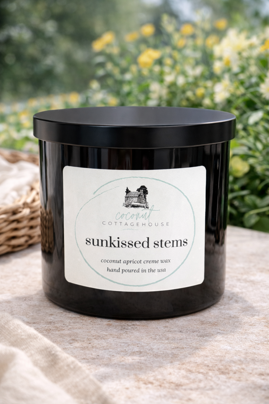 A black candle container with a label that reads "sunkissed stems" and "coconut apricot crème wax hand poured in the USA". The container is placed on a table with a wicker basket in the background.