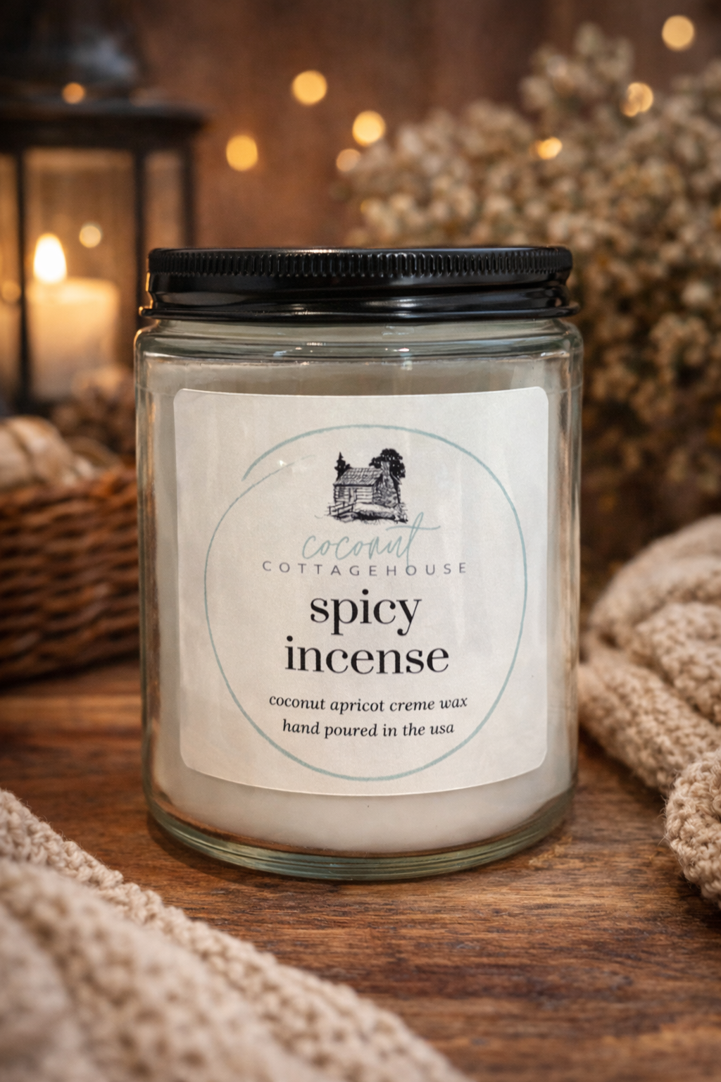 A glass jar candle labeled "spicy incense" from the "Cocoon Cottage" brand, with a wick and a black cap, sits on a wooden surface. The background includes a blurred image of a lantern and some greenery.