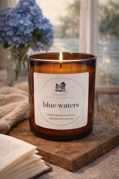 A brown candle labeled "blue waters" sits on a wooden surface, with a vase of blue hydrangeas in the background and an open book nearby.