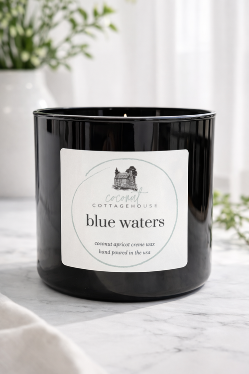 A black candle labeled "Blue Waters" from the "Cottage Cottage" brand, sitting on a white surface with a plant in the background.