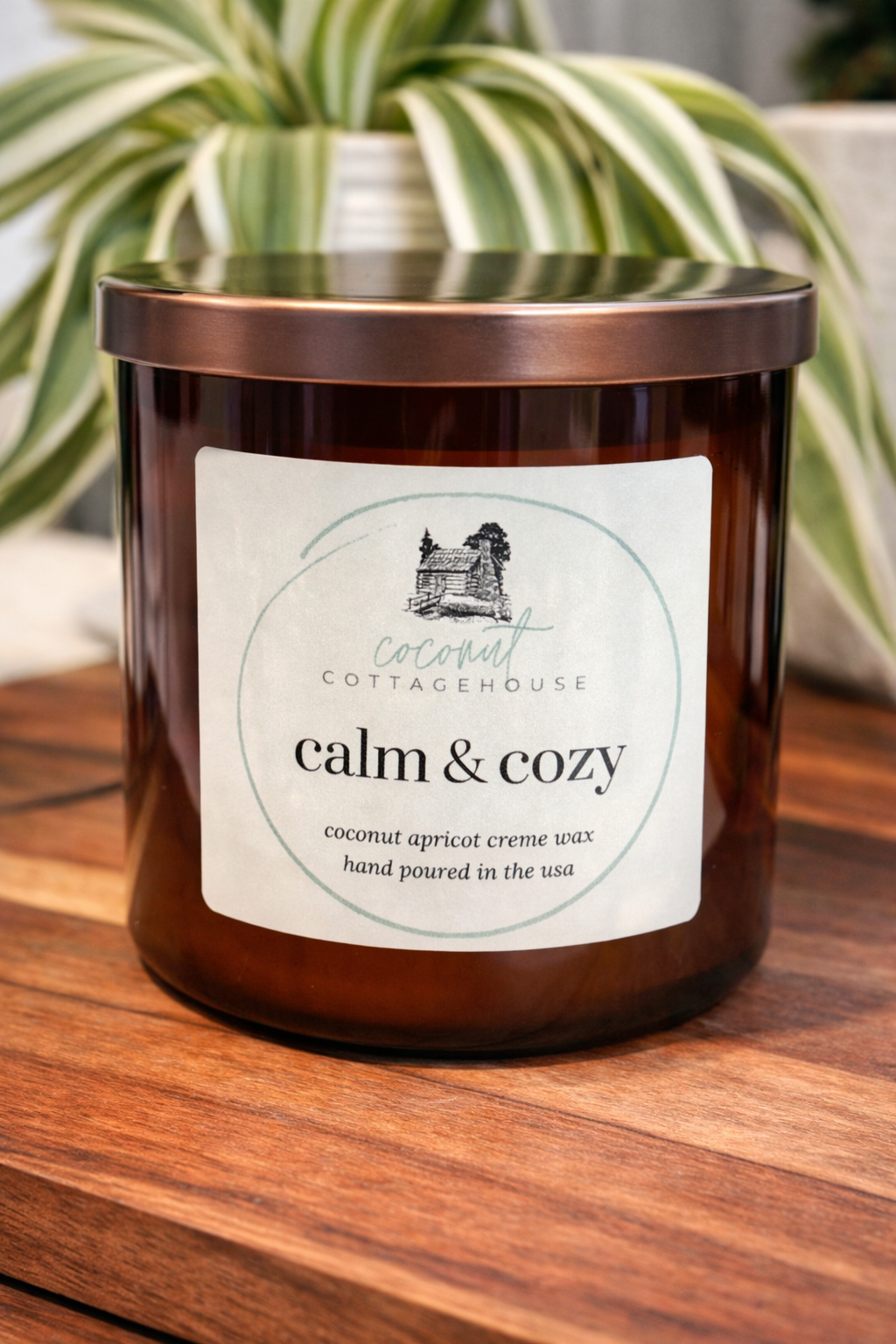 A brown glass candle labeled "Cottage Cottage" sits on a wooden surface, with a plant in the background.