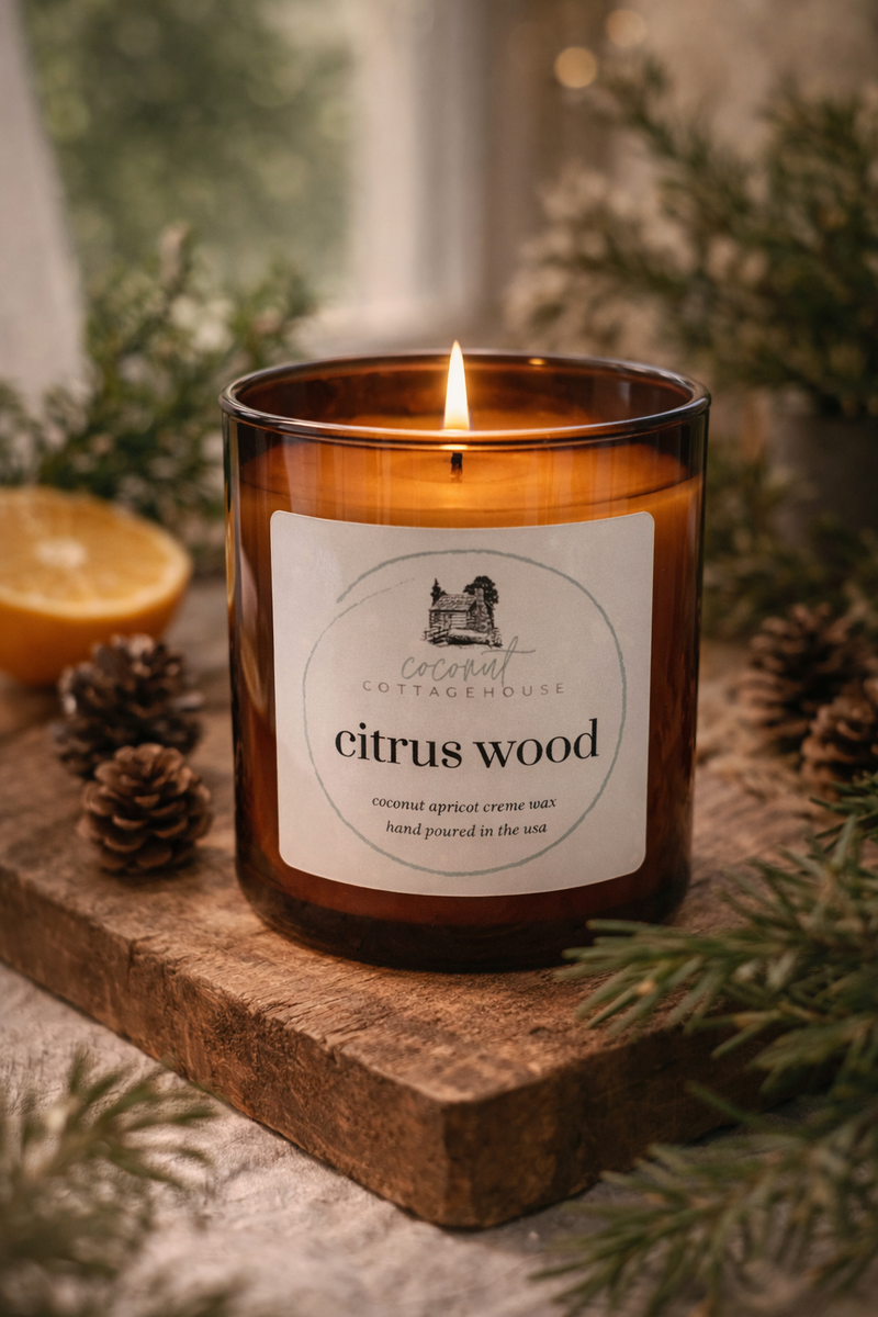 A brown candle labeled "Citrus Wood" sits on a wooden surface, surrounded by pine cones and an orange slice, creating a cozy and inviting atmosphere.