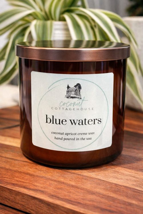 A brown glass candle labeled "Cottage Blue Waters" sits on a wooden surface, with a plant in the background.