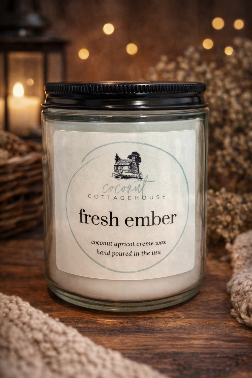 A glass jar candle labeled "fresh ember" from "Cocoon Cottage" sits on a wooden surface, with a blurred background featuring a lantern and twinkling lights.