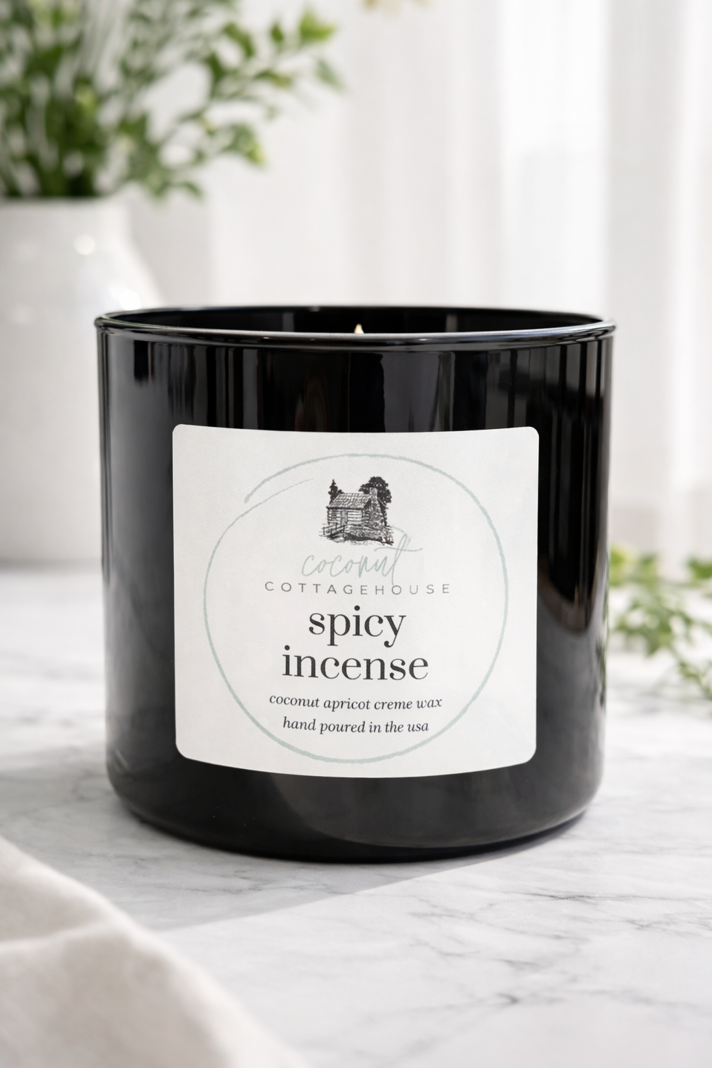 A black candle in a glass container with a label that reads "Coconut Cottage Spicy Incense".