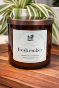 A brown glass candle labeled "fresh amber" from "Cottage Cottage" sits on a wooden surface, with a blurred plant in the background.