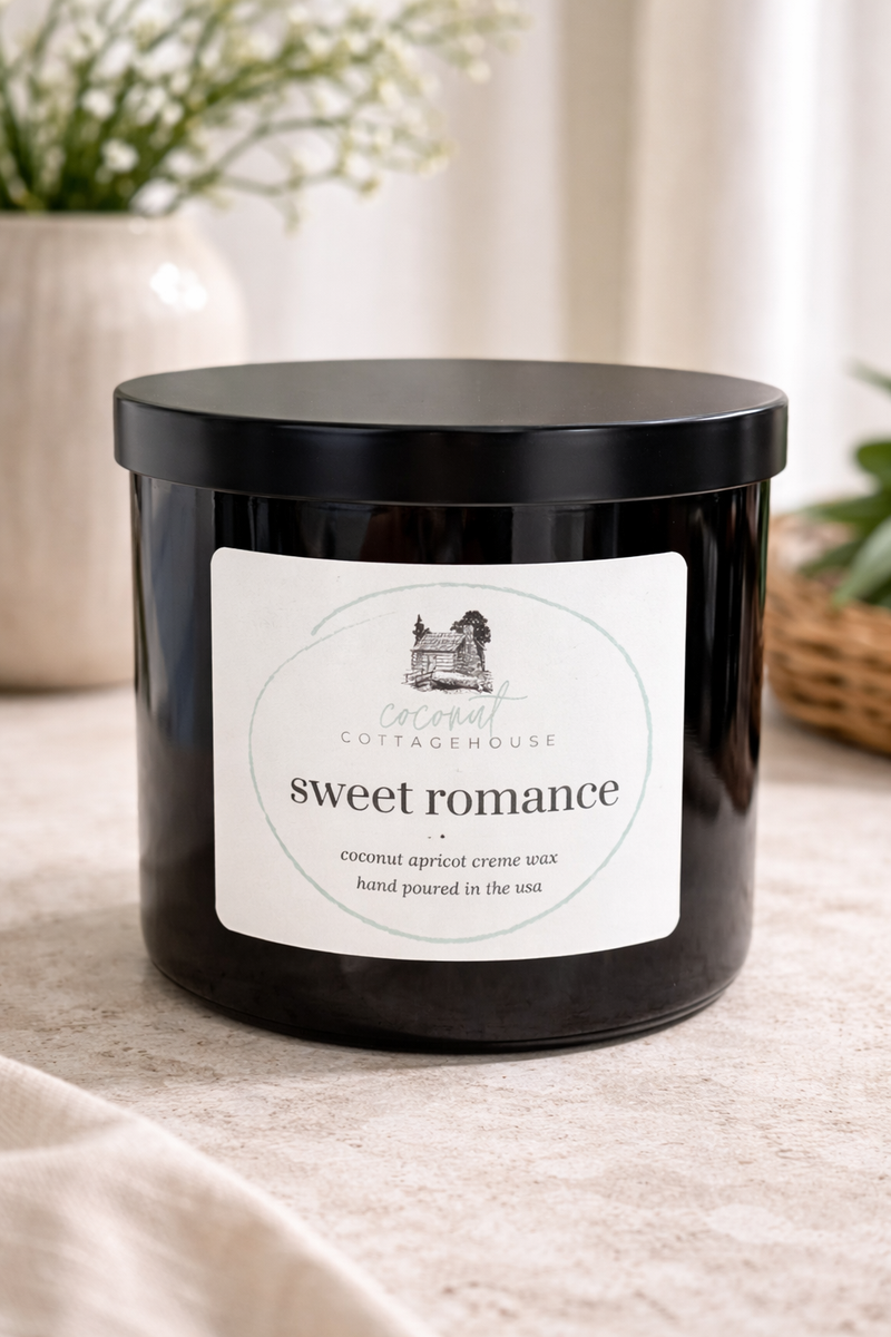 A black candle labeled "Sweet Romance" from the "Cottage Cottage" brand, sitting on a table with a wicker basket and a vase of flowers in the background.