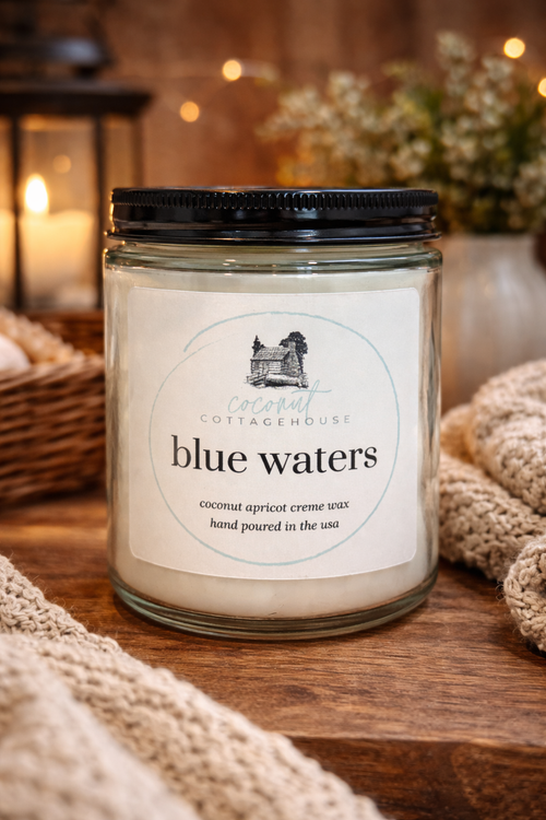 A glass jar candle labeled "Blue Waters" from the "Cottage Cottage" brand, sitting on a wooden surface with a blurred background that includes a lantern and a vase with flowers.