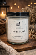 A glass jar candle labeled "Citrus Wood" from "Cottages" sits on a wooden surface, with a blurred background featuring a wicker basket and twigs.