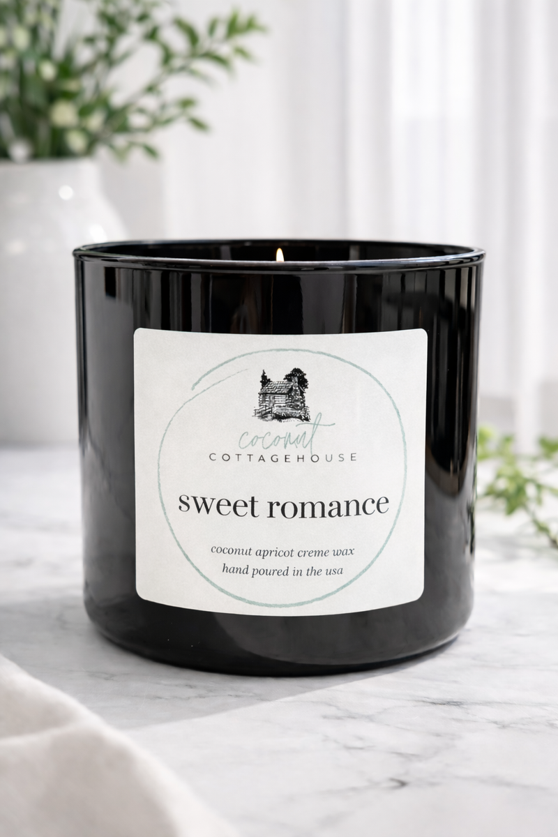 A black candle labeled "Sweet Romance" from the "Cottage" brand, sitting on a marble surface with a white vase of flowers in the background.