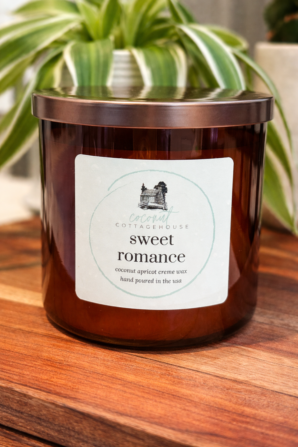 A brown glass candle with a label that reads "Sweet romance" sits on a wooden surface, with a plant in the background.