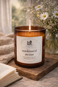 A brown glass candle labeled "sunkissed stems" sits on a wooden surface, with a vase of white flowers and a book nearby.