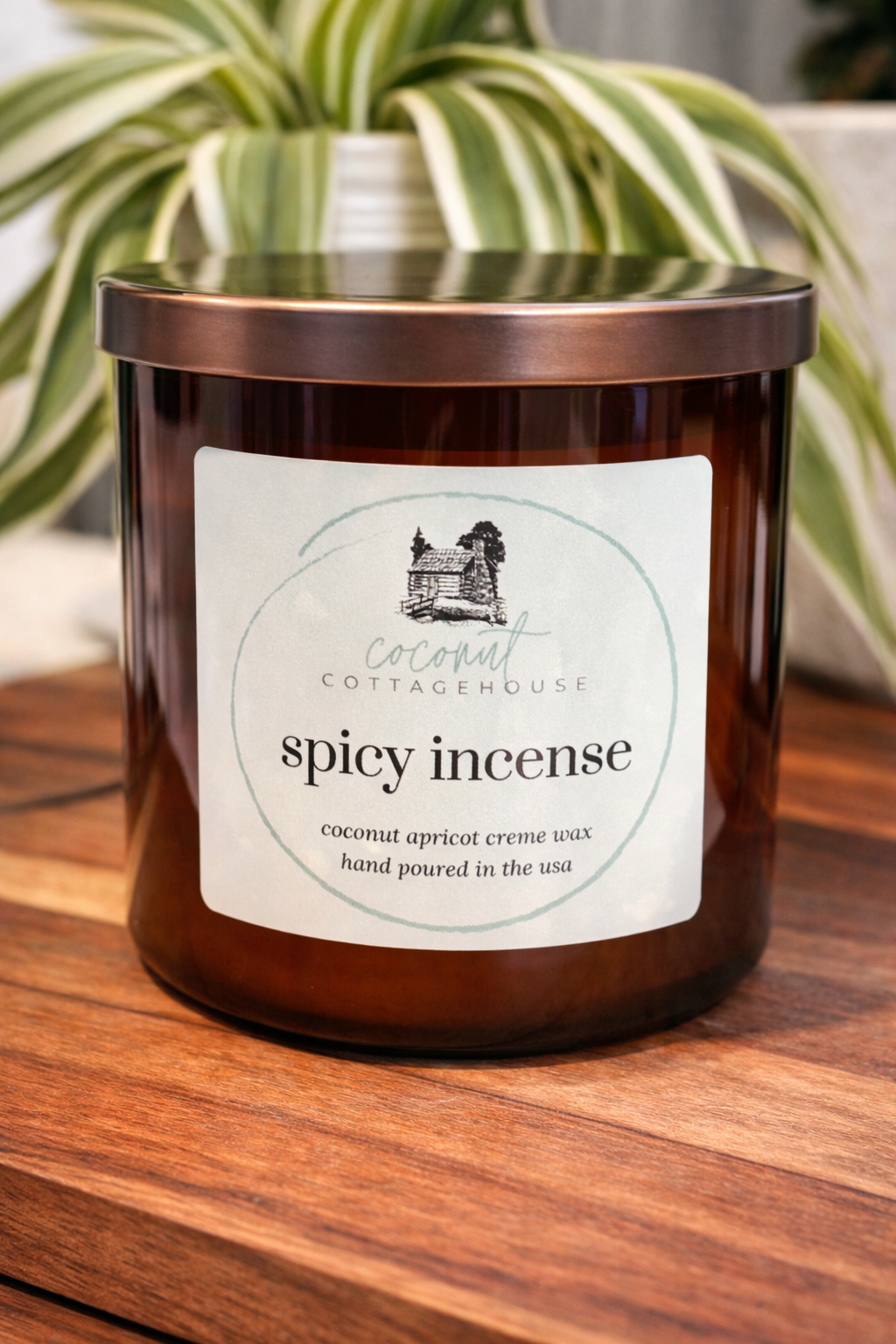 A brown glass candle labeled "Cottage Cottage" sits on a wooden surface, with a plant in the background.