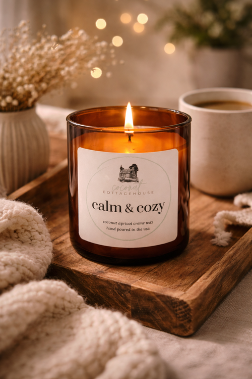 A brown glass candle labeled "calm & cozy" sits on a wooden tray, with a lit candle flame and a white mug of coffee nearby. The background is blurred, but it appears to be a cozy, warm setting with a vase of flowers and a string of lights.